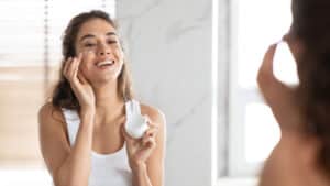 Happy young woman looking in mirror and applying medical-grade skin care product to face
